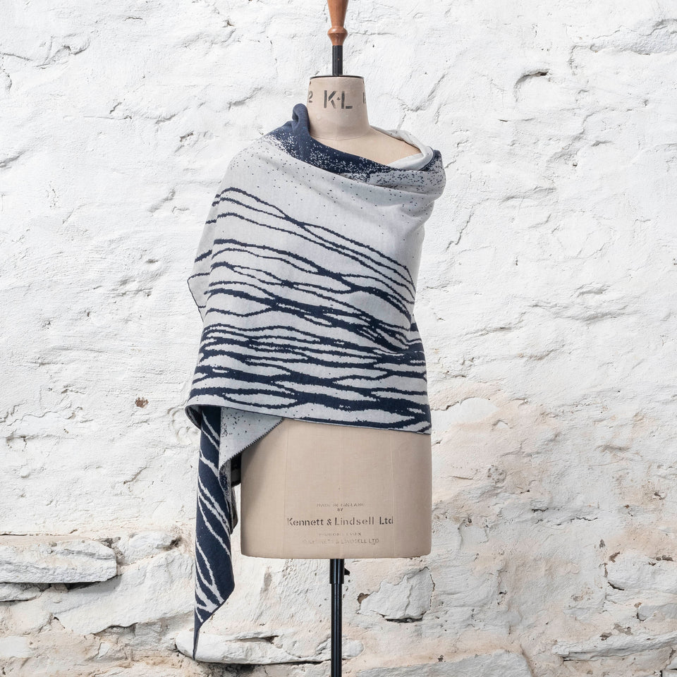 A midnight blue and off white knitted open cape in fine merino yarn. the pattern has fluid travelling likes either in blue on white or reversed out in white on blue. Shown from the front with linear pattern to the front wrapped round the mannequin