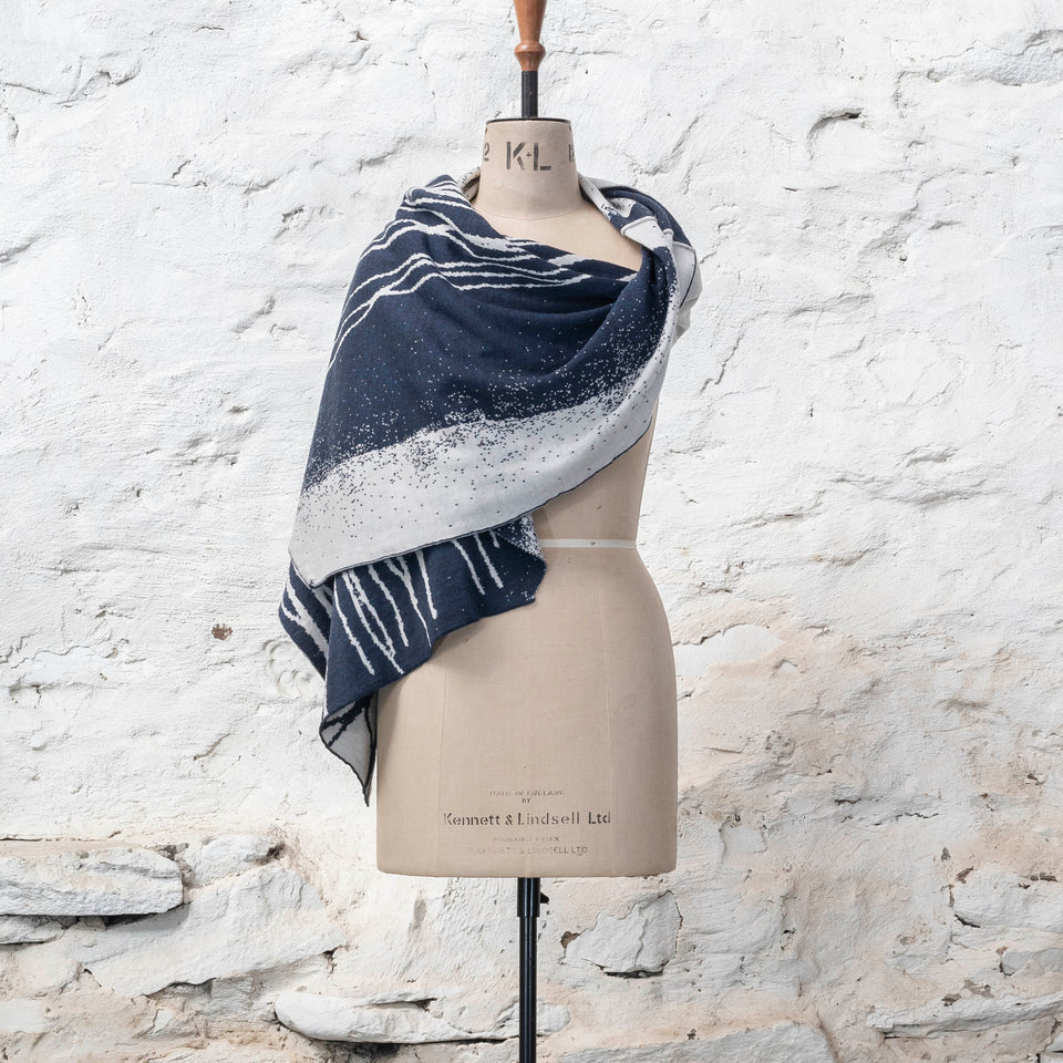 A midnight blue and off white knitted open cape in fine merino yarn. the pattern has fluid travelling likes either in blue on white or reversed out in white on blue. Shown from the front in a sweeping wrap to the right shoulder