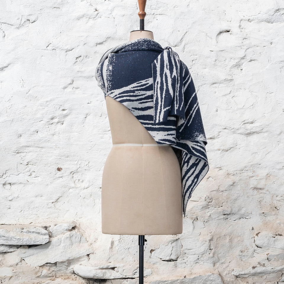 A midnight blue and off white knitted open cape in fine merino yarn. the pattern has fluid travelling likes either in blue on white or reversed out in white on blue. Shown from back sweeping over the right shoulder