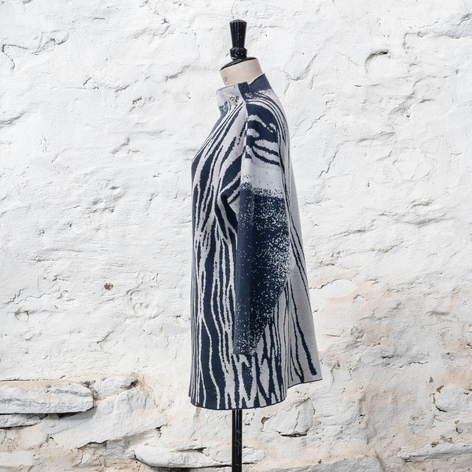 On a vintage mannequin, shown against a rustic whitewashed wall, a longline tunic jumper finely knitted in midnight blue and off white. Shown from the back right with lines travelling down over the shoulders to the bottom of the garment. The tunic has a stand-up neck