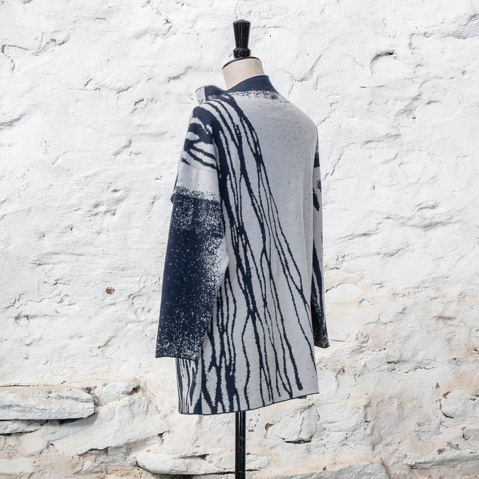 On a vintage mannequin, shown against a rustic whitewashed wall, a longline tunic jumper finely knitted in midnight blue and off white. Shown from the back right in a three quarters view, where the pattern is predominantly white with blue lines travelling from the left shoulder down to the bottom of the garment. The tunic has a stand-up neck