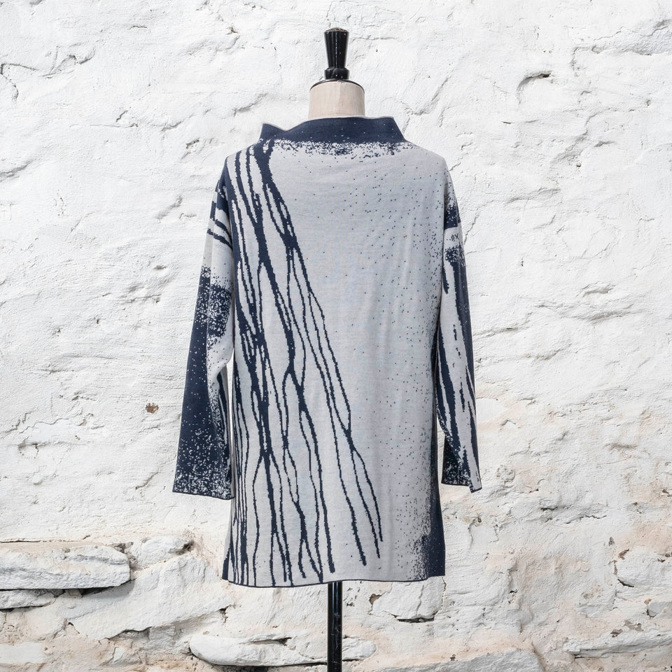 On a vintage mannequin, shown against a rustic whitewashed wall, a longline tunic jumper finely knitted in midnight blue and off white. Shown from the back where the pattern is predominantly white with blue lines travelling from the right shoulder down to the bottom of the garment. The tunic has a stand-up neck