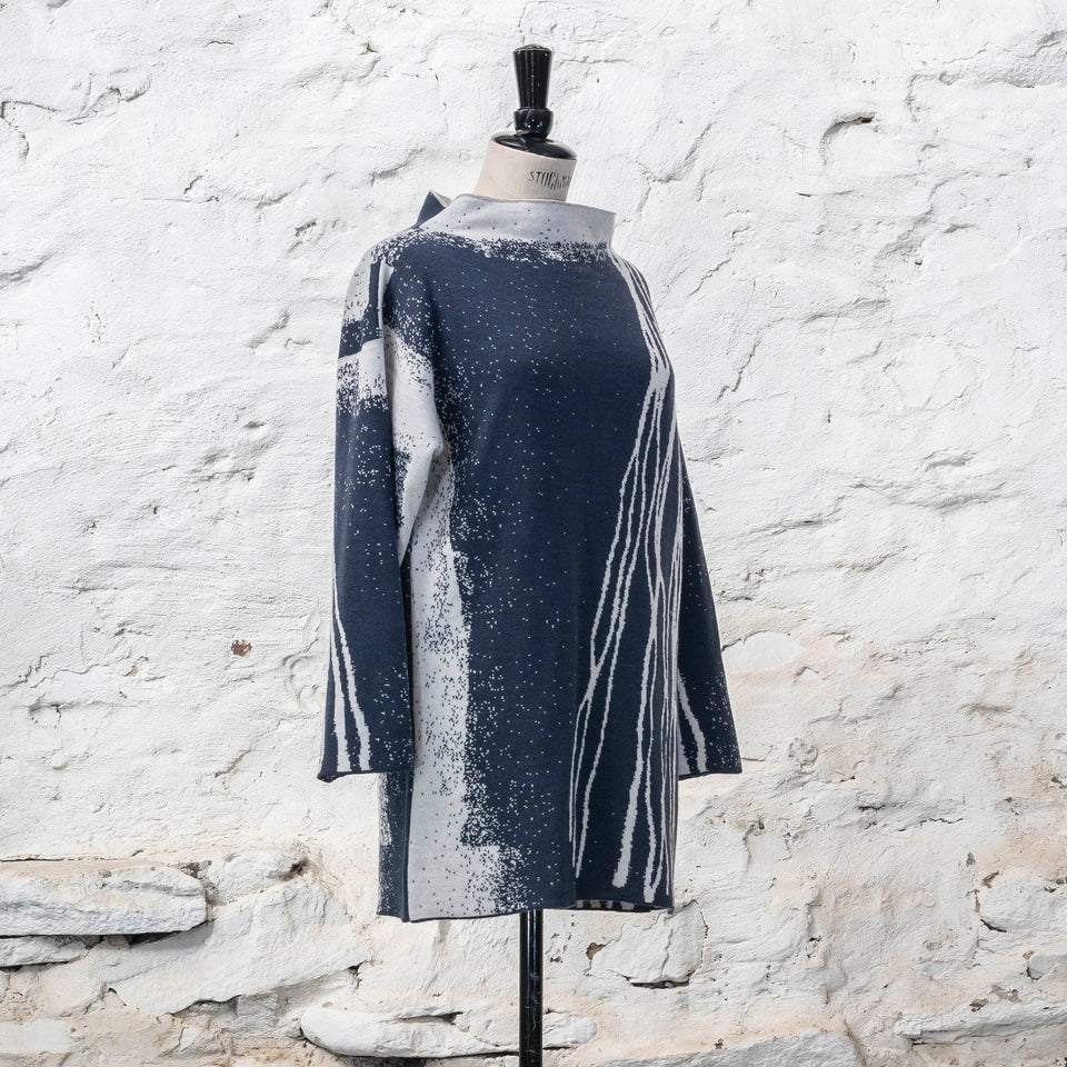 On a vintage mannequin, shown against a rustic whitewashed wall, a longline tunic jumper finely knitted in midnight blue and off white. Shown with the predominantly blue area to the front, withy white with blue lines travelling from the left shoulder down to the bottom of the garment. The tunic has a stand-up neck