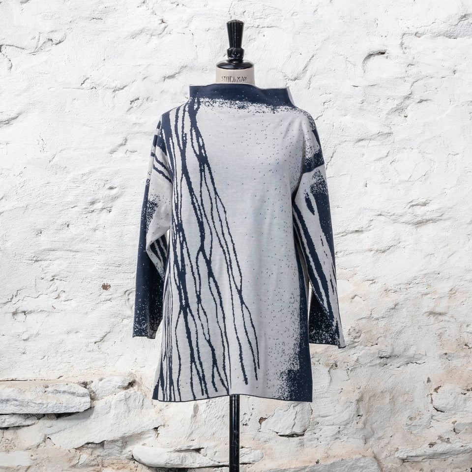 On a vintage mannequin, shown against a rustic whitewashed wall, a longline tunic jumper finely knitted in midnight blue and off white. Shown from the front where the pattern is predominantly white with blue lines travelling from the right shoulder down to the bottom of the garment. The tunic has a stand-up neck 