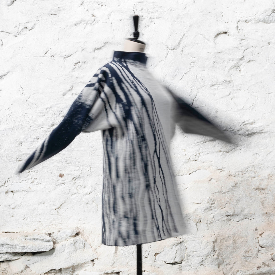 On a vintage mannequin, shown against a rustic whitewashed wall, a longline tunic jumper finely knitted in midnight blue and off white. Shown from the back right in a three quarters view, where the pattern is predominantly white with blue lines travelling from the right shoulder down to the bottom of the garment. The tunic has a stand-up neck. The sleeves fly up as the manequin is twisted