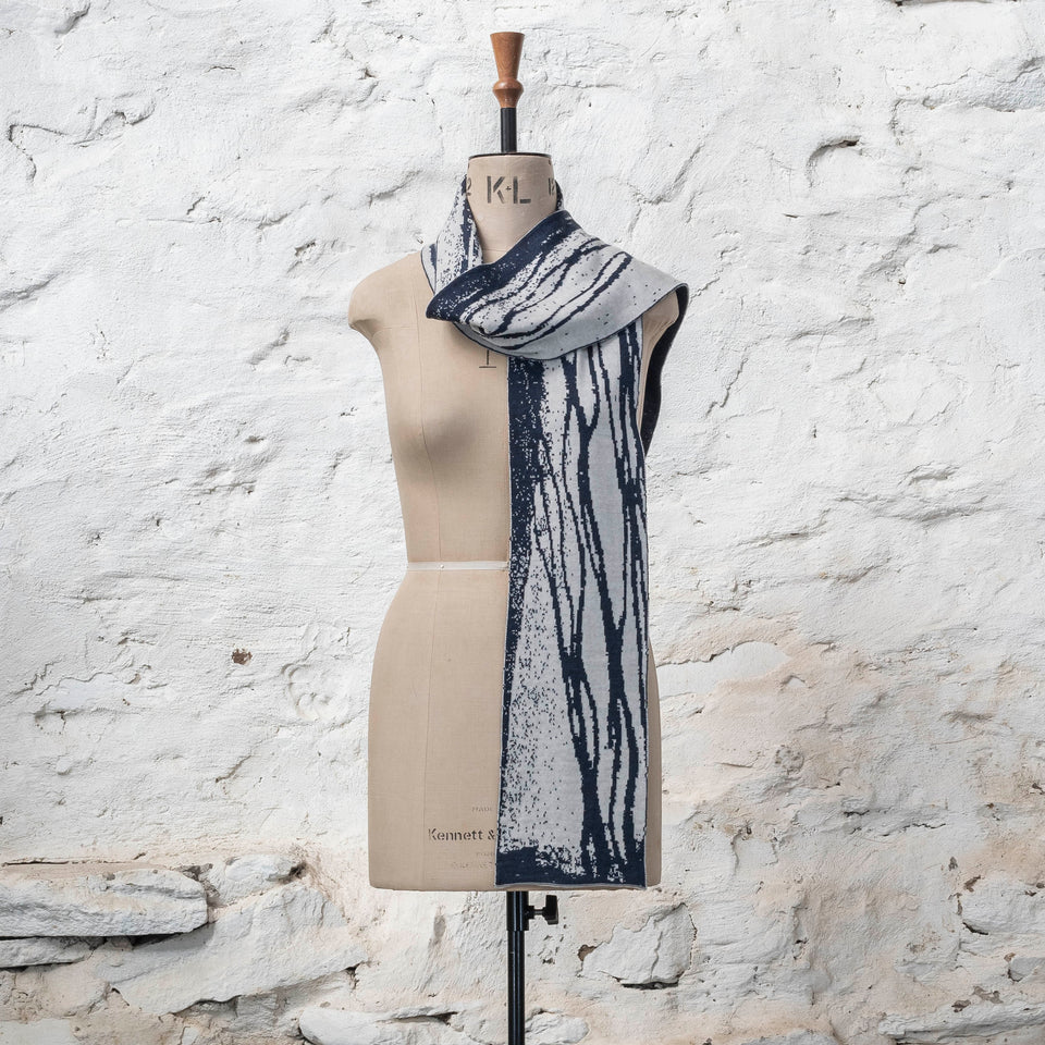 On a vintage mannequin shown against a rusting whitewashed wall, a midnight blue and off white finely knitted scarf in reverse jacquard. The pattern is linear wavs of line and speckle, moving along the scarf. Shown with one end thrown over the shoulder, the other hanging down the front.