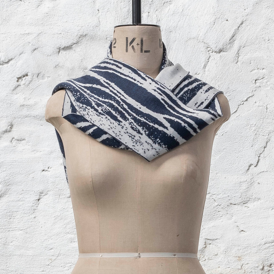 On a vintage mannequin shown against a rusting whitewashed wall, a midnight blue and off white finely knitted scarf in reverse jacquard. The pattern is linear wavs of line and speckle, moving along the scarf. Shown with ends of scarf thrown over shoulder.