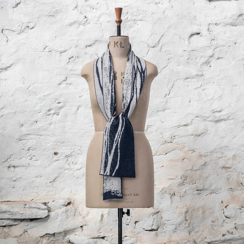 On a vintage mannequin shown against a rusting whitewashed wall, a midnight blue and off white finely knitted scarf in reverse jacquard. The pattern is linear wavs of line and speckle, moving along the scarf. Shown in a loose knot at the waist.