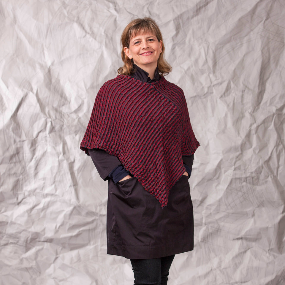 Model wears rigg cape. The textile is ridged and with stripes of colour knitted in. Shown here in a dark red and navy