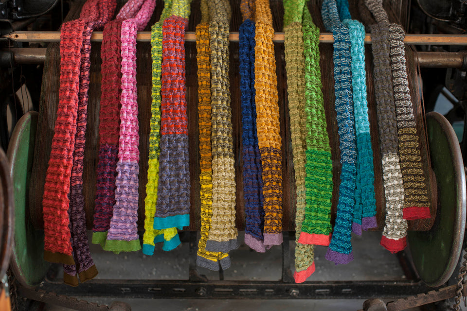 Selection of rigg scarves arranged on an old loom. The scarves are in a ridged textile, with stripes and coloured blocks. The ends are a different stripe