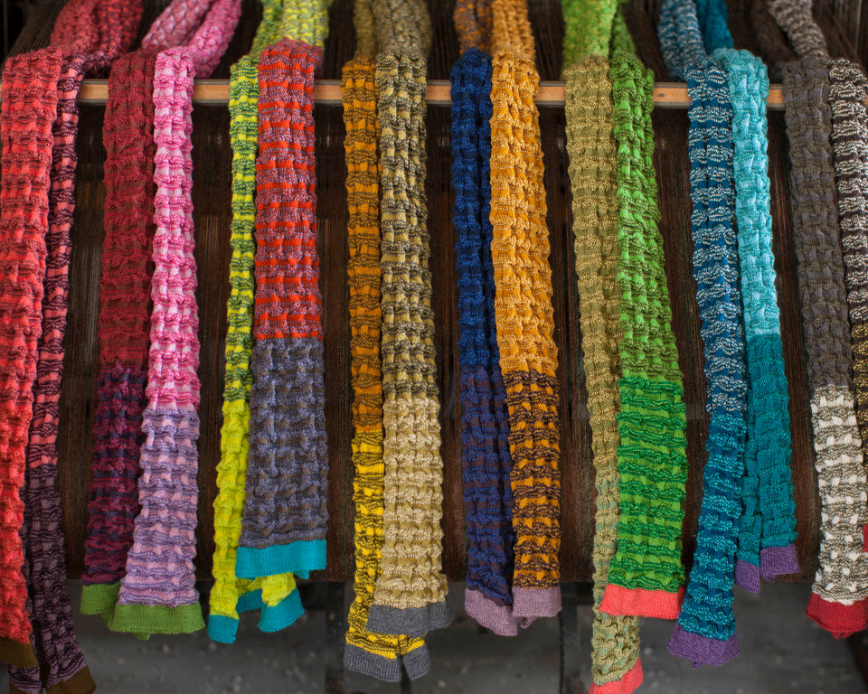Selection of rigg scarves arranged on an old loom. The scarves are in a ridged textile, with stripes and coloured blocks. The ends are a different stripe