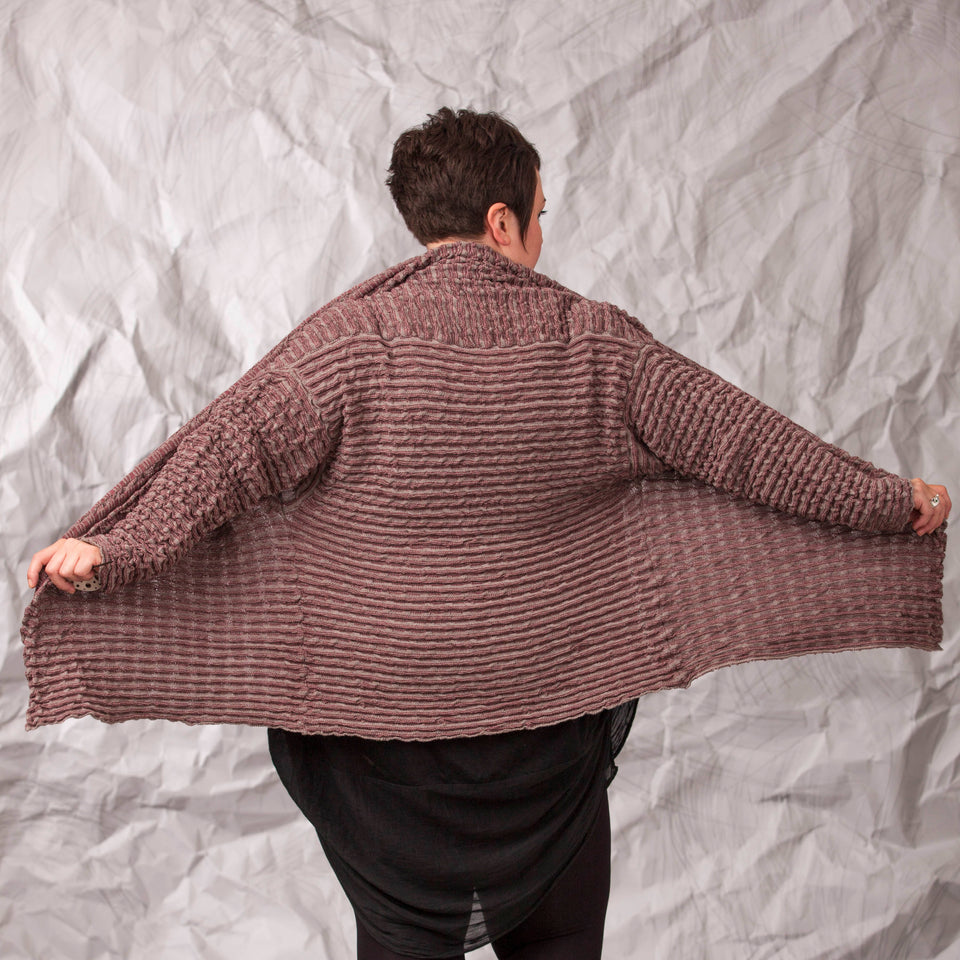 Model wears a longline knitted contemporary jacked in ridged fabric, in warm beige and cherry. Back view with front of jacket opened out to show shape