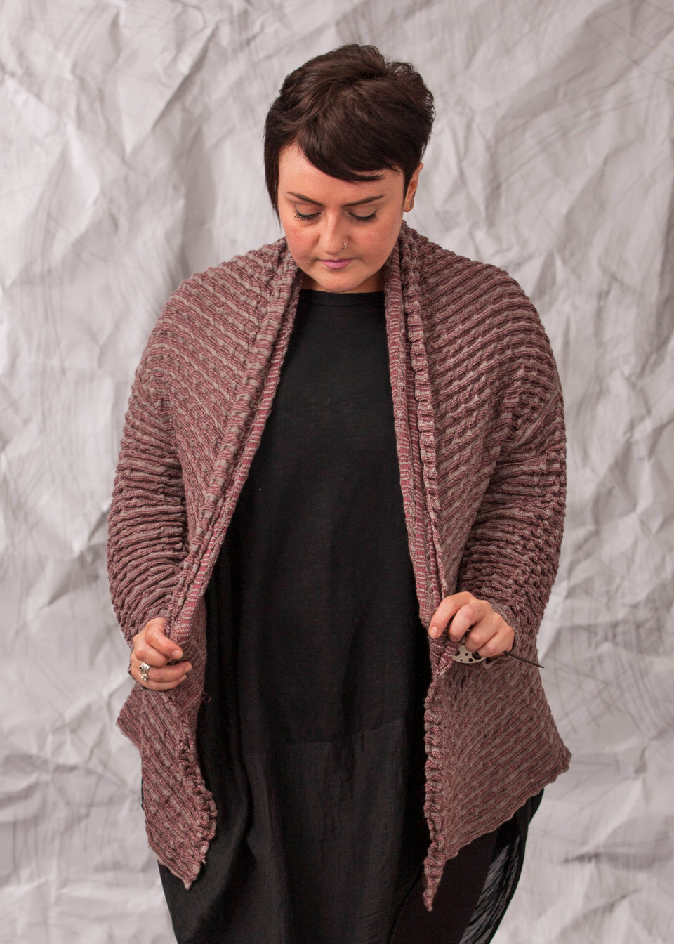 Model wears a longline knitted contemporary jacked in ridged fabric, in warm beige and cherry. Front view, holding the jacket slightly open.