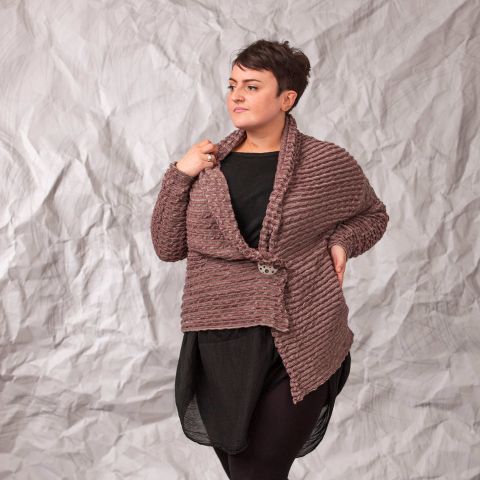 Model wears a longline knitted contemporary jacked in ridged fabric, in warm beige and cherry. Three-quarter view, with the jacket pinned shut