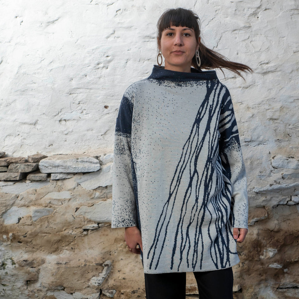 A woman with long dark hair and olive skin looks towards the camera. She is wearing cream metal hoop earrings and her he hair tied back in a ponytail, which swishes to one side. She is wearing a longline tunic jumper in navy and off white. The design is abstract and linera with large areas of speckled pattern. She is also wearing black trousers and is standing in a stone, whitewashed shed in hoswick Shetland.