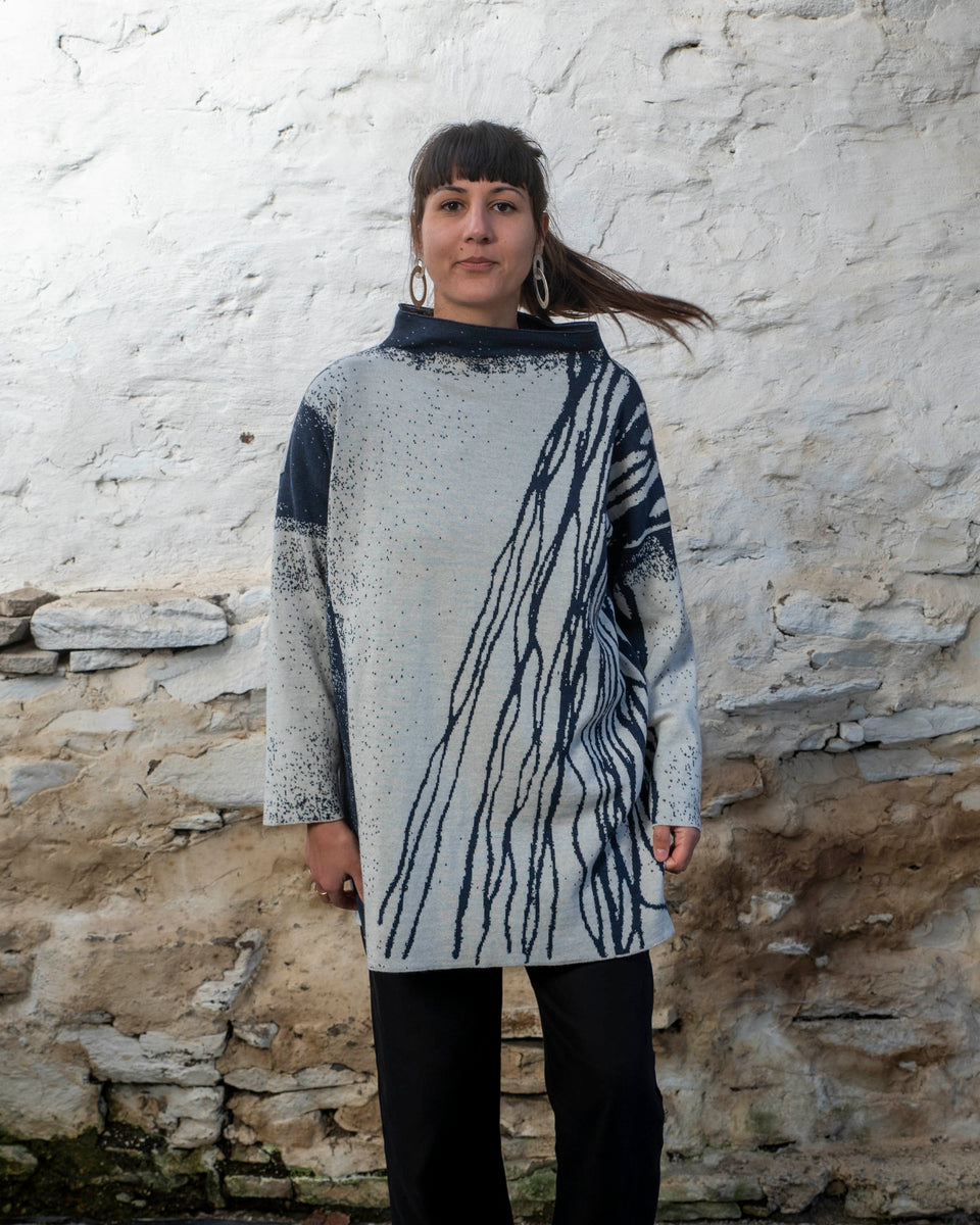 A woman with long dark hair and olive skin looks towards the camera. She is wearing cream metal hoop earrings and her he hair tied back in a ponytail, which swishes to one side. She is wearing a longline tunic jumper in navy and off white. The design is abstract and linera with large areas of speckled pattern. She is also wearing black trousers and is standing in a stone, whitewashed shed in hoswick Shetland.