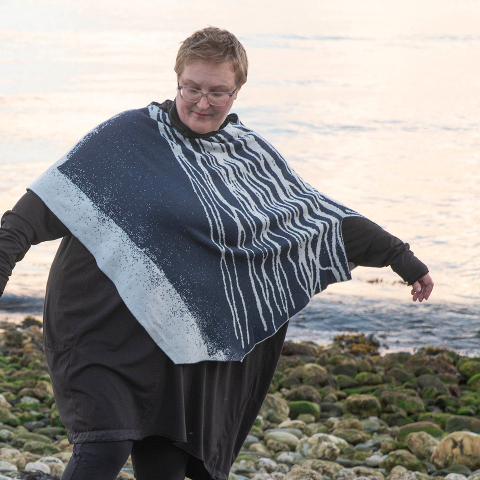 A white woman with short fair hair stands on a pebbly beach in Hoswick Shetland. She is wearing purplish clear spectacles, an off black rundholz tunic and leggings. Over this she wears a finely knitted cape by Nielanell.  The design is abstract with flowing lines reminiscent of water rivulets on sand and is knitted in inky blue and off white reverse jacquard.