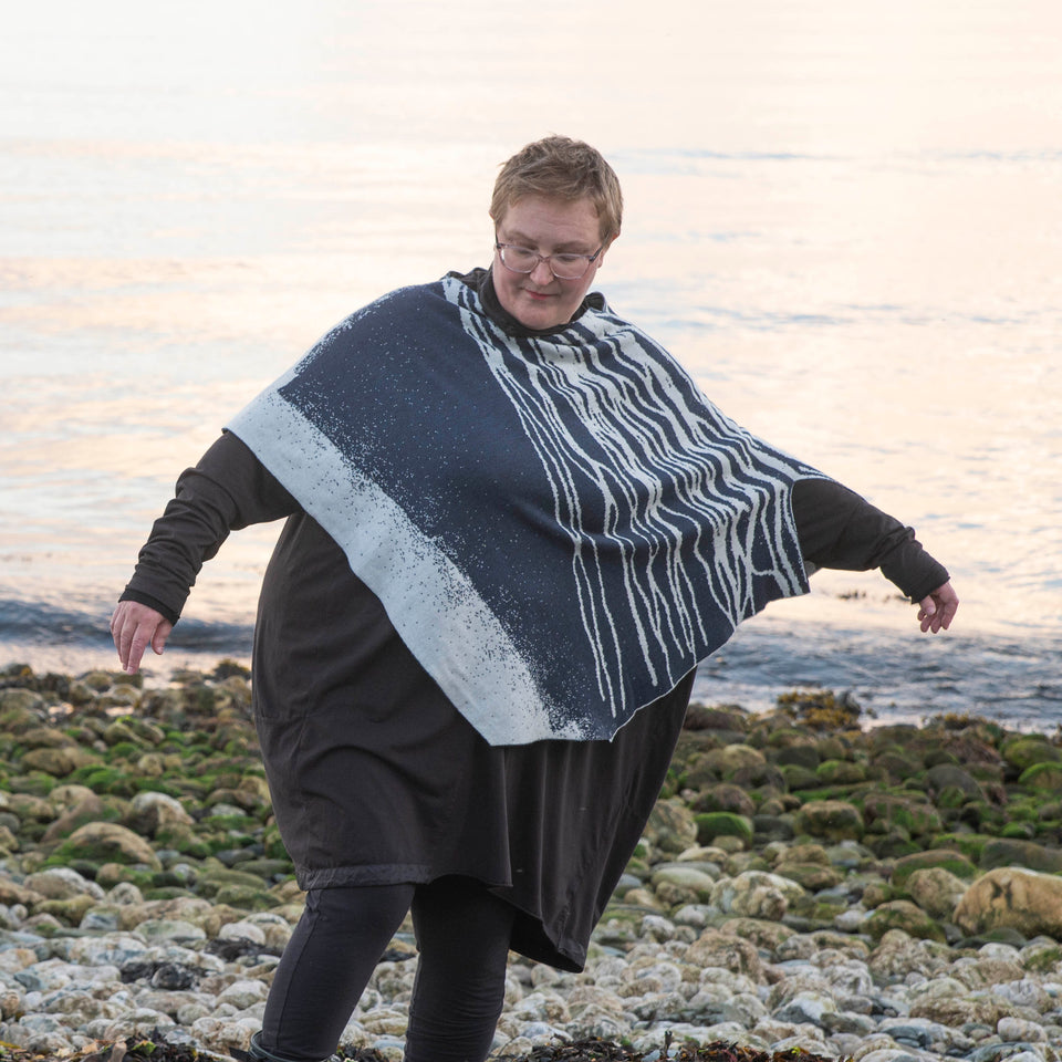 A white woman with short fair hair stands on a pebbly beach in Hoswick Shetland. She is wearing purplish clear spectacles, an off black rundholz tunic and leggings. Over this she wears a finely knitted cape by Nielanell.  The design is abstract with flowing lines reminiscent of water rivulets on sand and is knitted in inky blue and off white reverse jacquard.