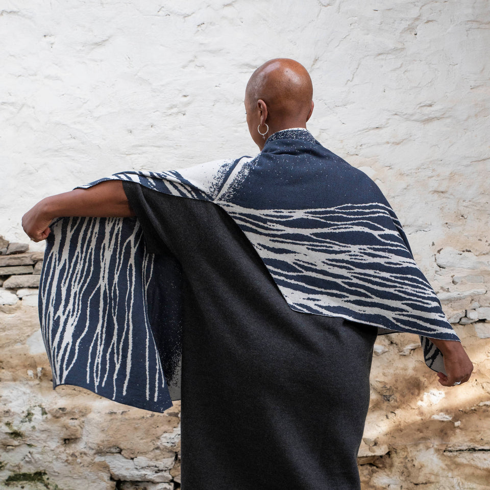 A black woman with a shaved head stands in a rustic, whitewashed Shetland building. She is wearing a charcoal grey woollen dress. Over this, she wears a contemporary wrap in inky blue and off white. The wrap as pattern of linear flowing lines, whcih are shown as she lifts her left arm (she has her back to the camera). The wrap has a fish tail shape with a V notch taken out at the back.