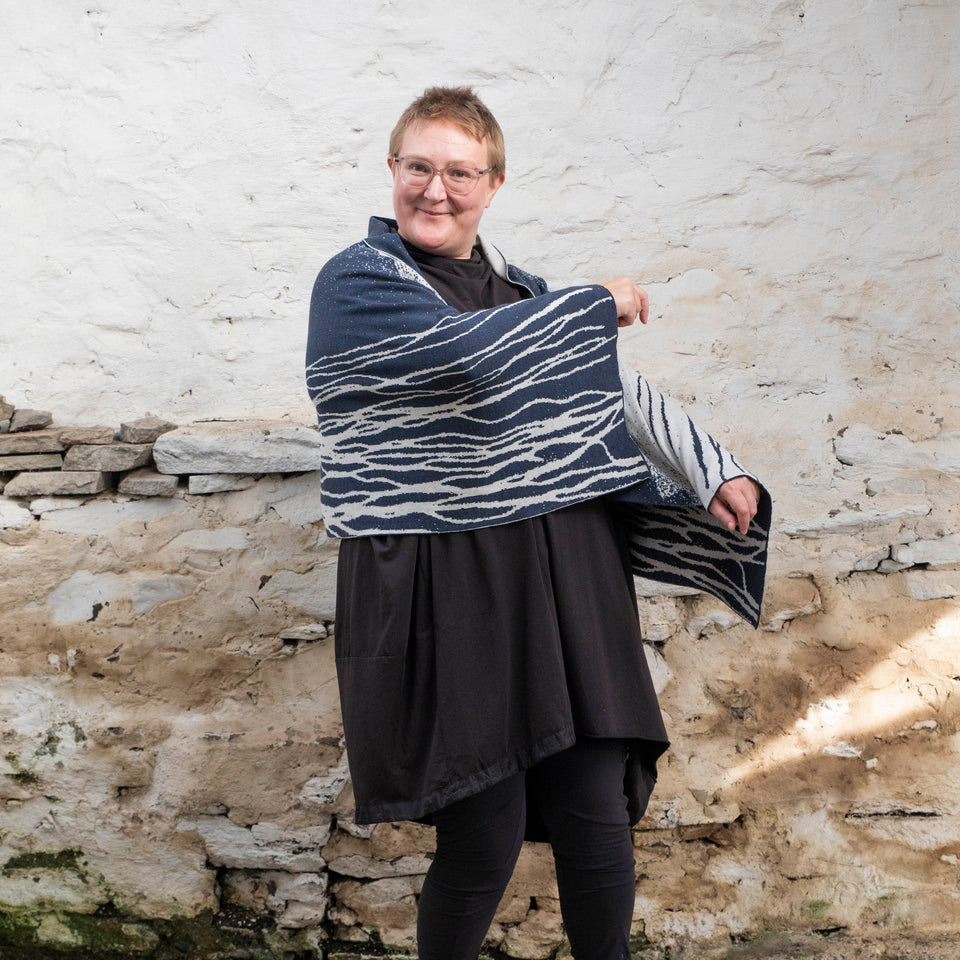 A white woman with short, fair hair stands with her left side to the camera in a rustic, whitewashed Shetland stone shed. She is wearing spectacles with thin metal legs. Over an off-black tunic she wears a contemporary, finely knitted shawl. A pattern of flowing lines travel along her arms. She brings her right arm up to shoulder height, showing the pattern. The shawl is dark inky blue and off-white. She is looking towards the camera 